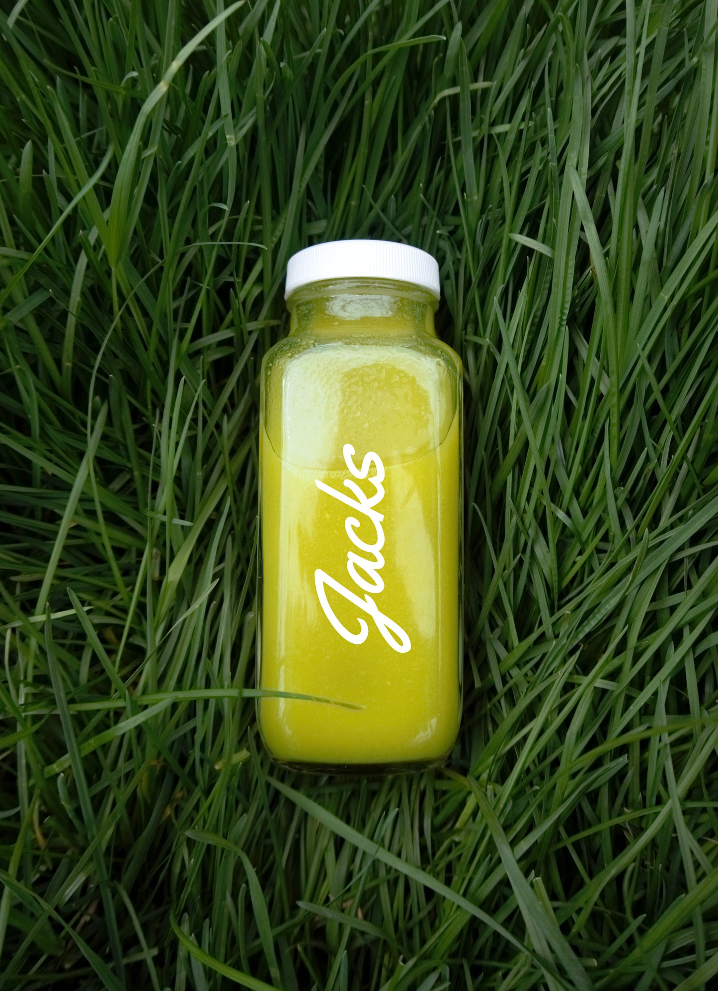 A 250ml glass bottle of light green liquid, with a simple "Jacks" written in white cursive font placed vertically up the length of the bottle, nestled in a background of lush green grass.