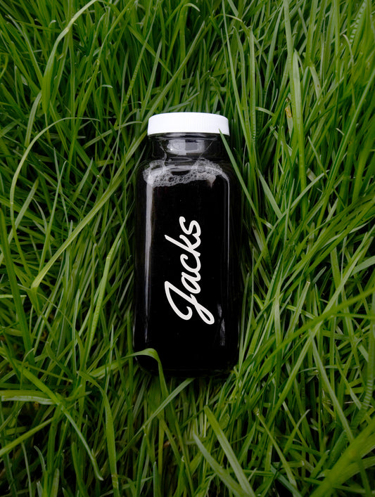 A 250ml glass bottle of deep black liquid, with a simple "Jacks" written in white cursive font placed vertically up the length of the bottle,  nestled in a background of lush green grass.